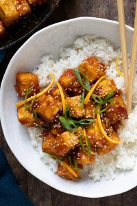 Orange chicken sprinkled with sesame seeds in large white bowl over white rice