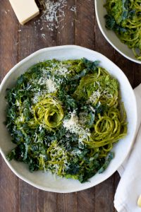 white bowl filled with creamy pesto pasta swirls, topped with kale and parmesan cheese on wooden table