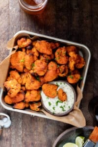 baked cauliflower buffalo wings in square container with a glass of cold beer and limes