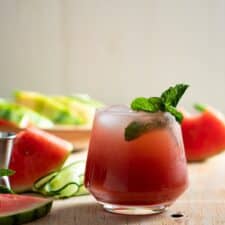 cocktail on light wood board with watermelon slices and cucumber ribbons beside it