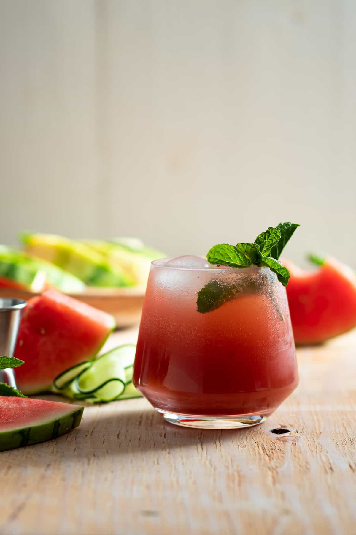 cocktail on light wood board with watermelon slices and cucumber ribbons beside it