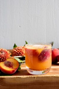 bourbon peach cocktail on wood table with white background near peach colored flowers
