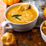 white bowl filled with pumpkin soup with sage leaves and a piece of bread dunked in