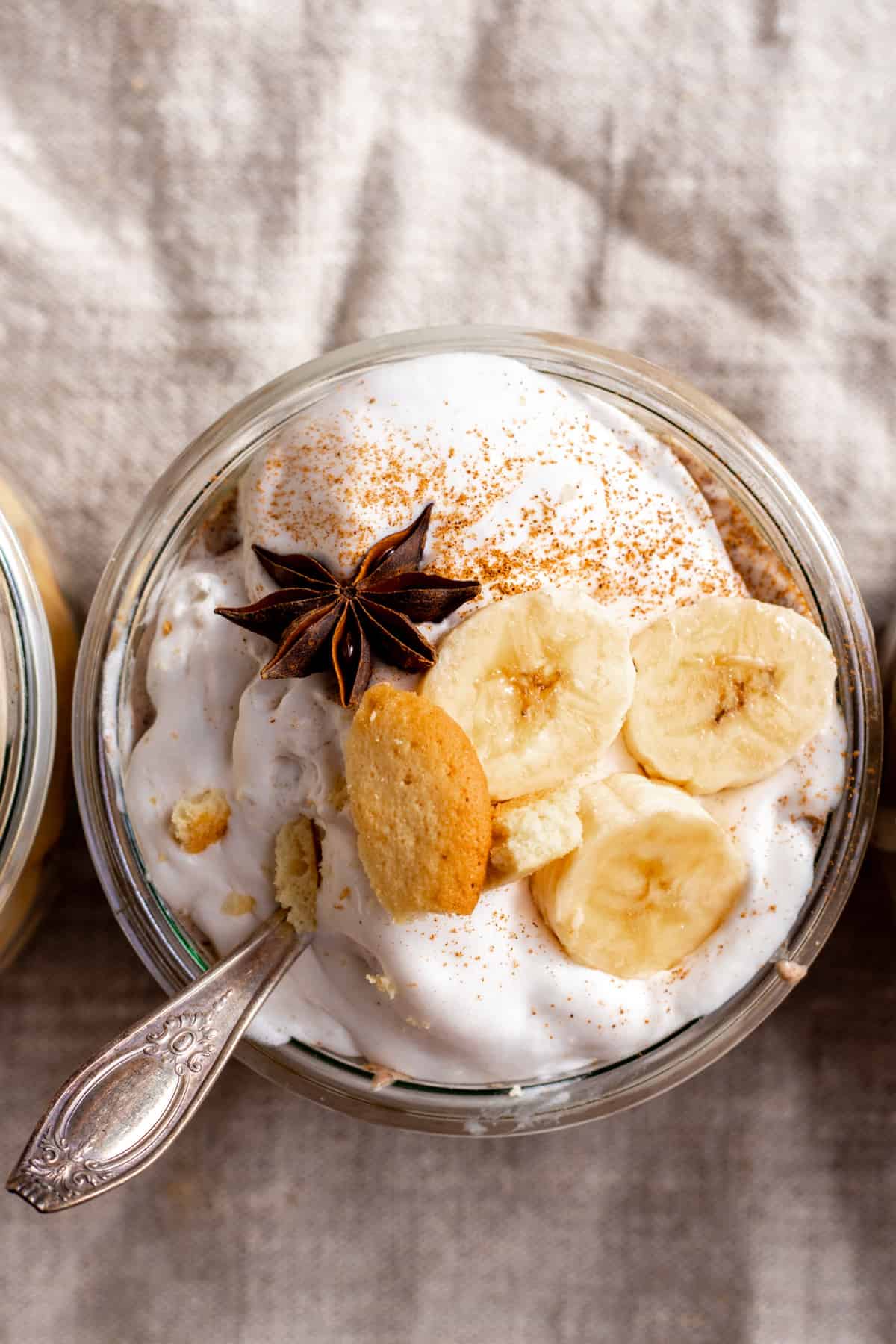 Sliced banana and star anise on top of pudding with spoon in jar.