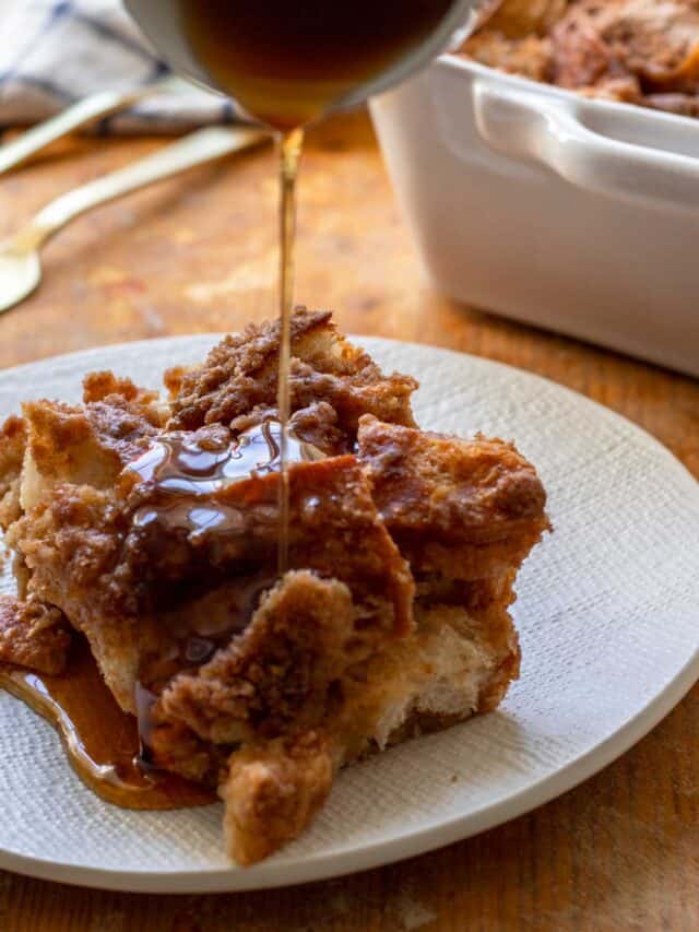 Slice of french toast casserole on white plate with maple syrup being poured over top.