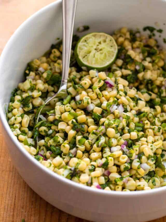 Corn salsa with half lime in large white bowl and silver spoon.