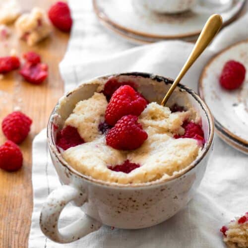 Vanilla mug cake topped with fresh raspberries and a gold spoon.