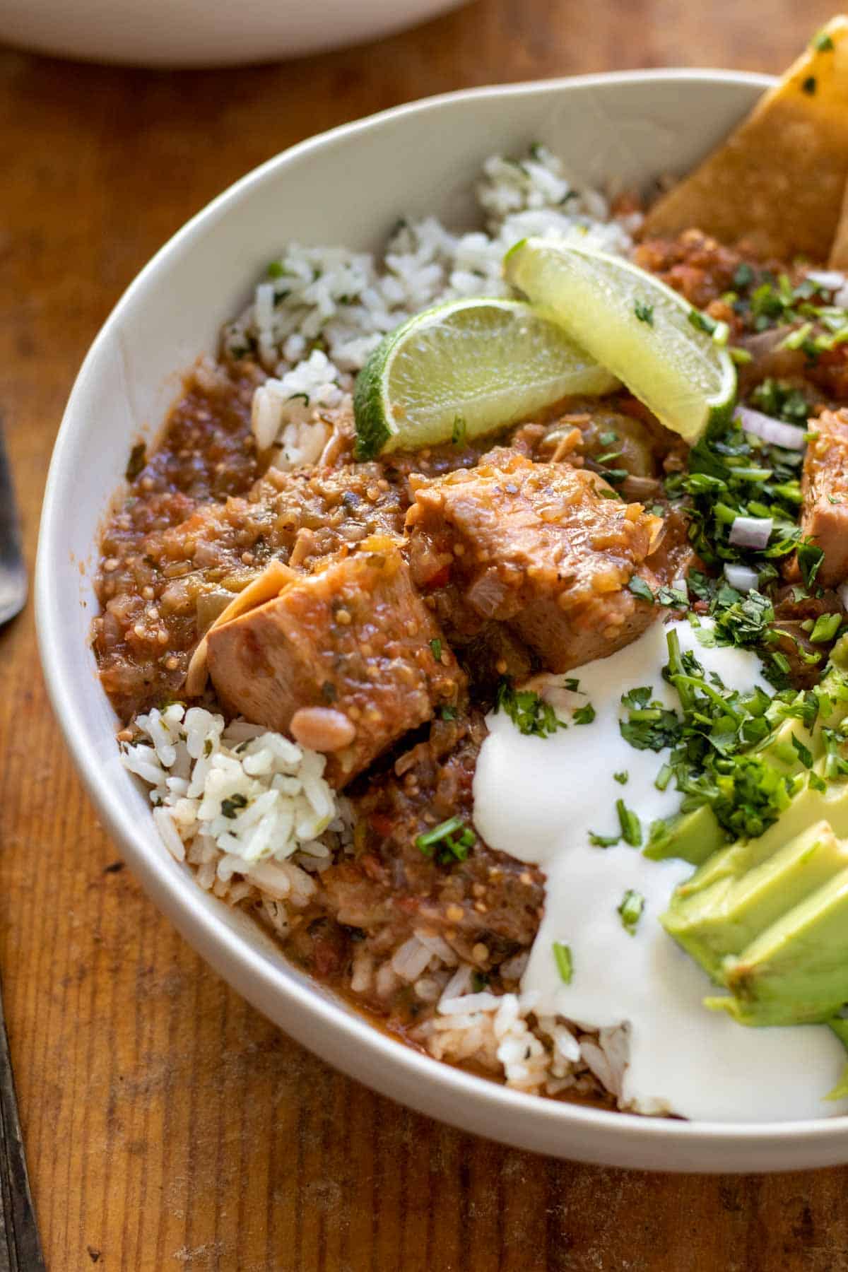 Close up of stew served over rice in white bowl topped with avocado slices, sour cream, cilantro, and tortilla chips.