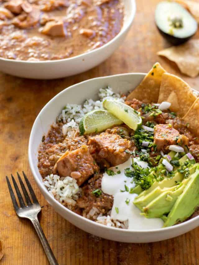 Stew topped with sour cream, avocado slices, corn chips over cilantro rice in white bowl.