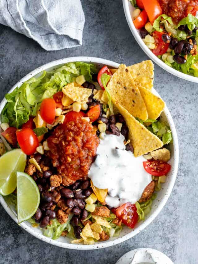 Taco salad bowl garnished with salsa and lime wedges in white bowl.