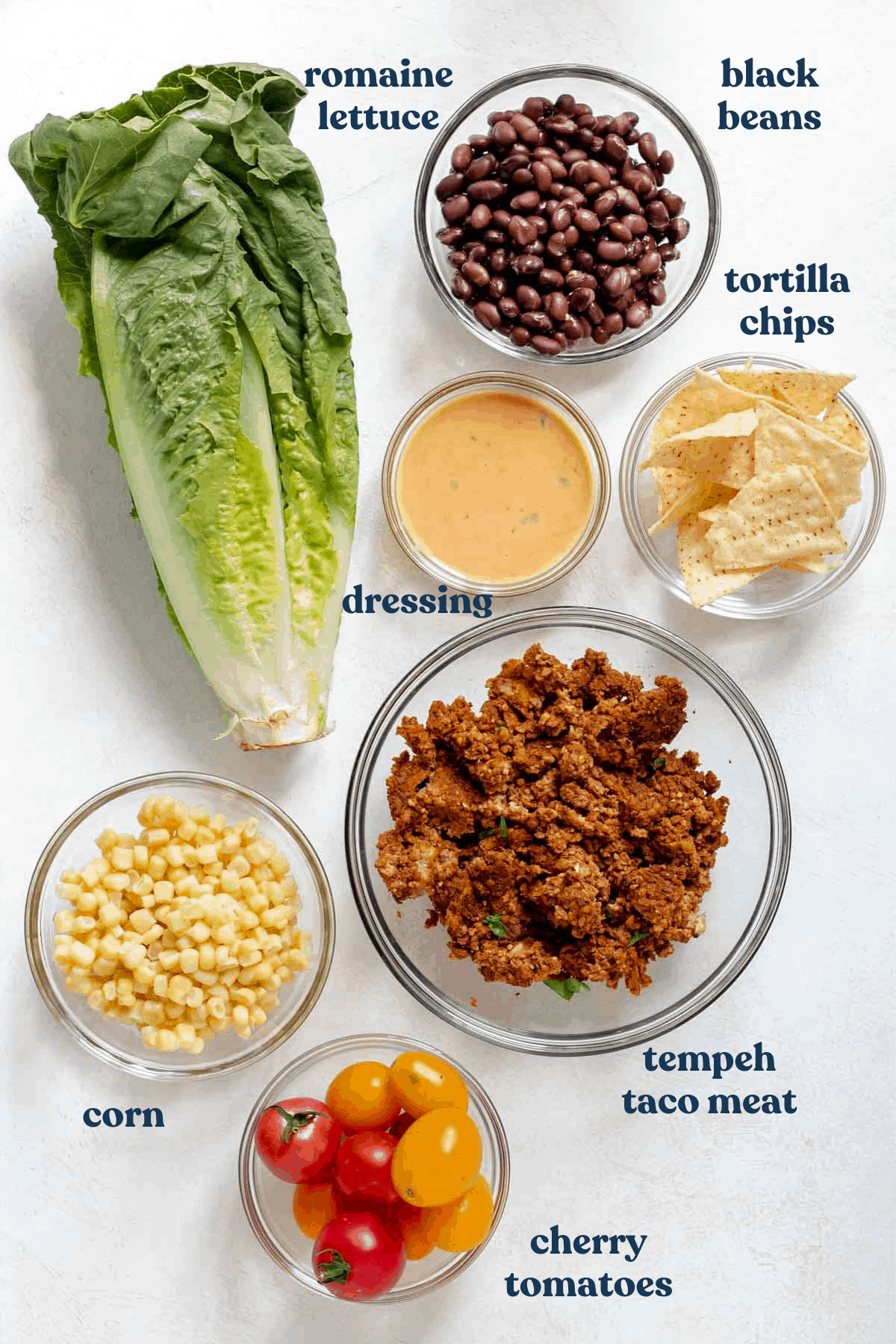 Black beans, romaine lettuce, tortilla chips, dressing, taco meat, corn and tomatoes on white backdrop.
