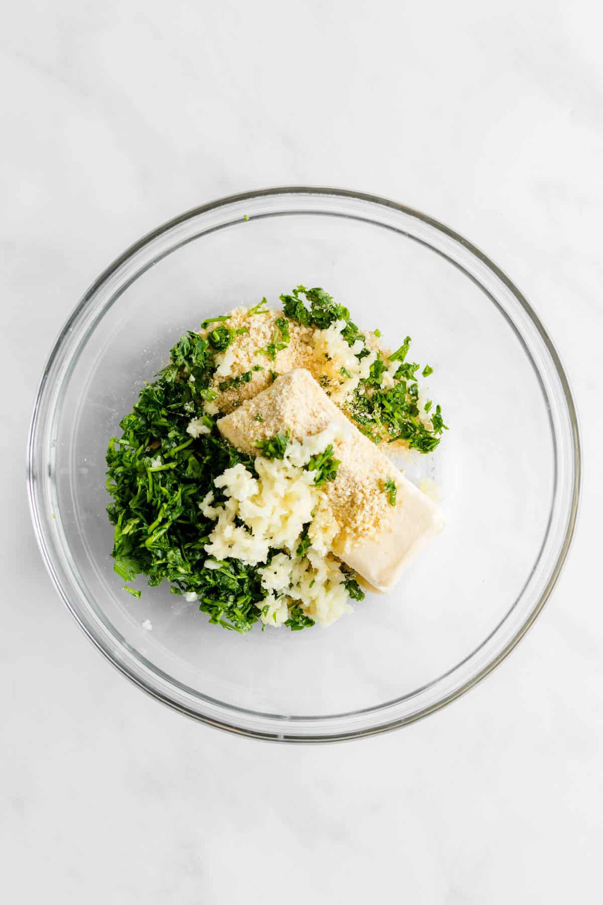 Softened butter, minced garlic, parsley, and vegan parmesan in a glass mixing bowl.