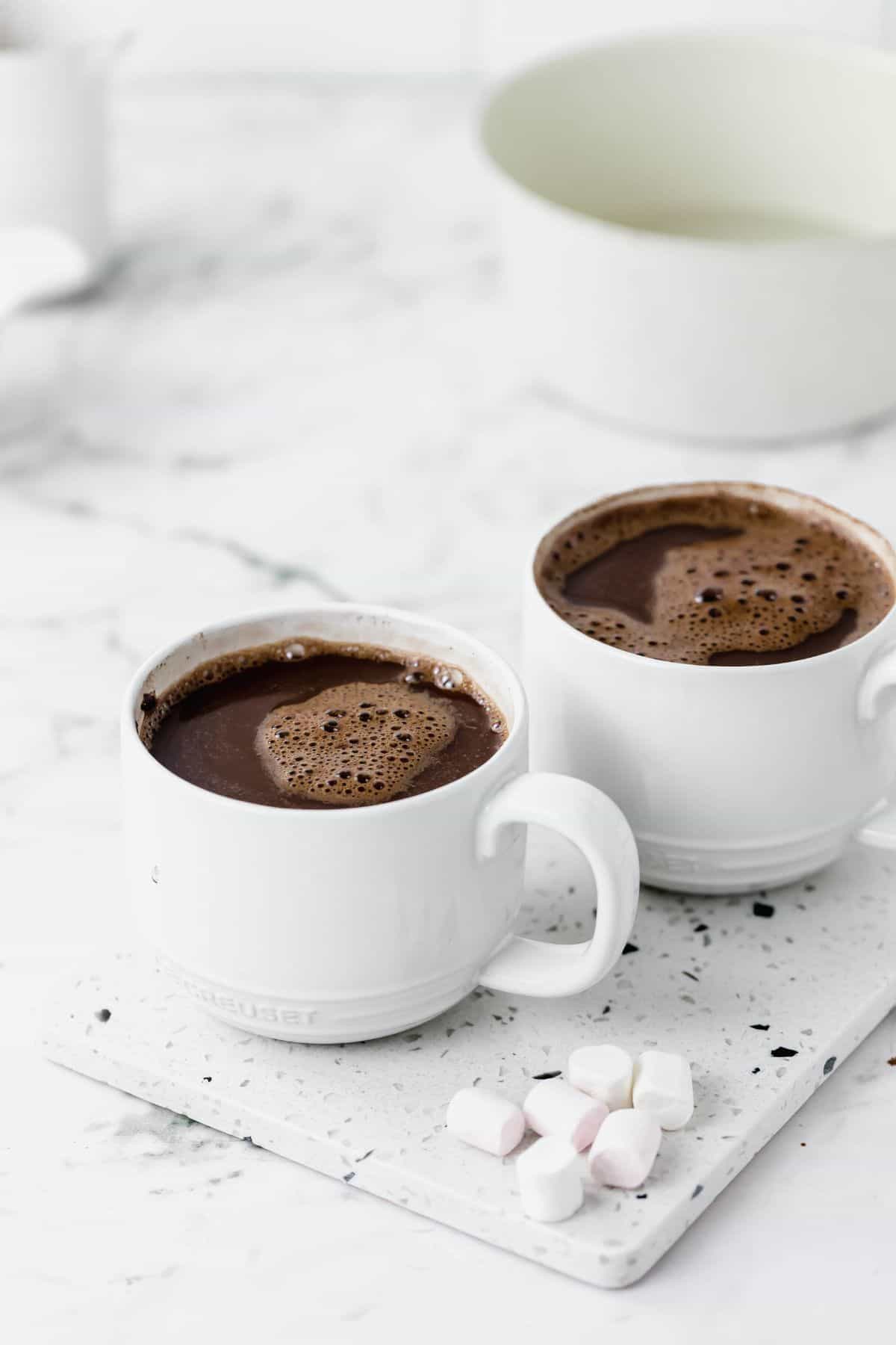 Two white mugs of cocoa by marshmallows.