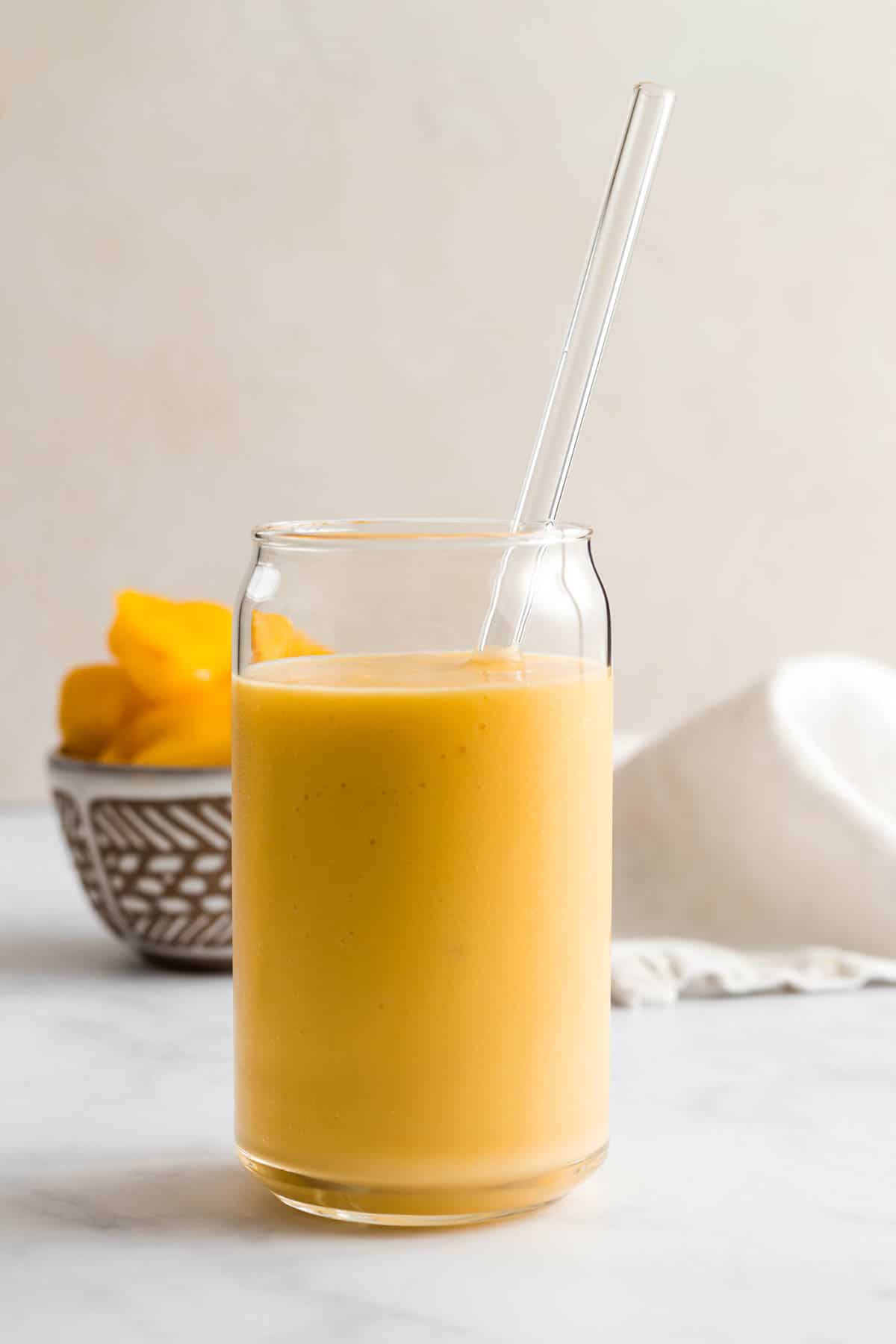 A yellow smoothie in glass cup with glass straw.