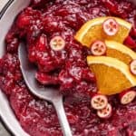 Closeup of homemade bourbon cranberry sauce in serving bowl with fresh cranberries and orange slices garnish.
