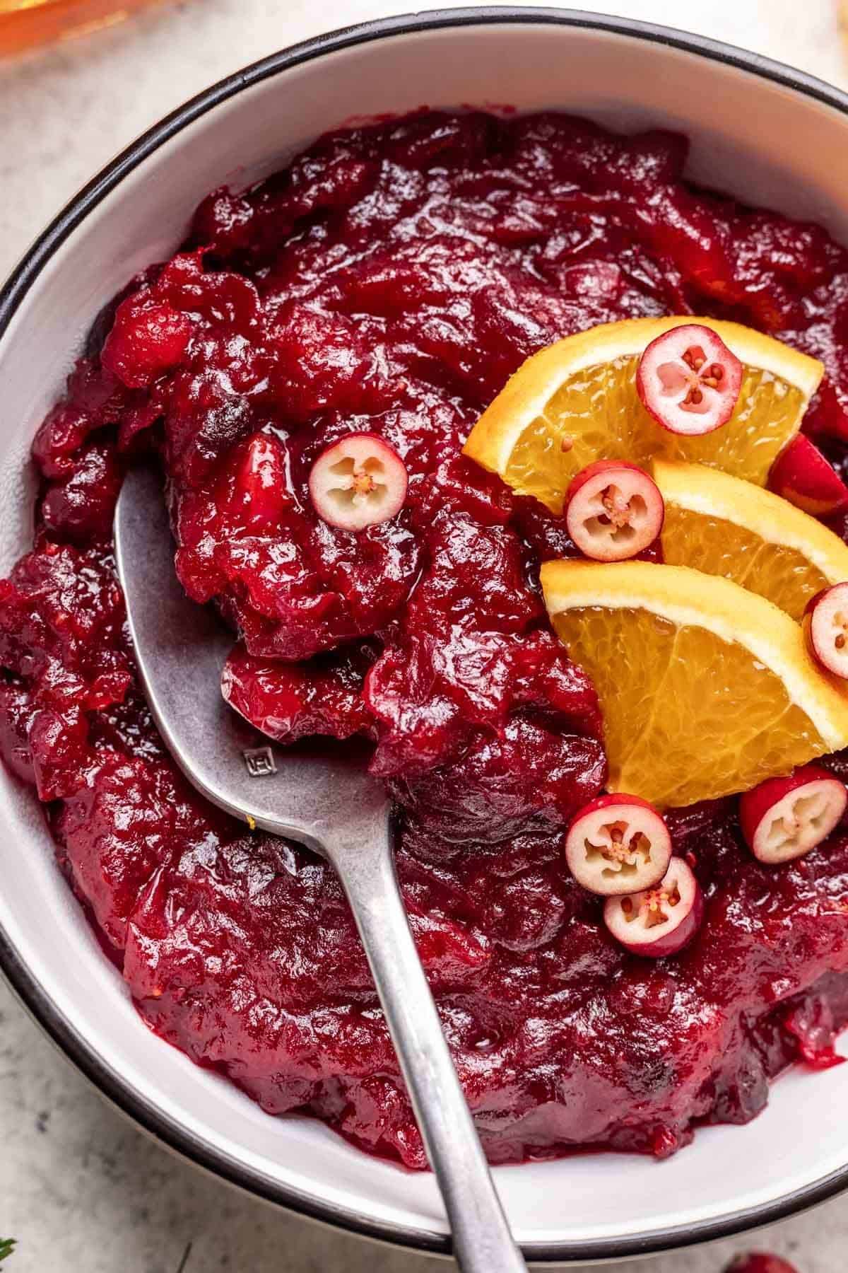 Closeup of homemade bourbon cranberry sauce in serving bowl with fresh cranberries and orange slices garnish.