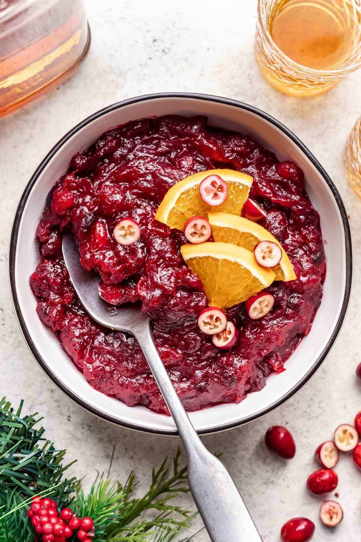 Bowl of cranberry sauce with a serving spoon in bowl and garnished with fresh orange slices.